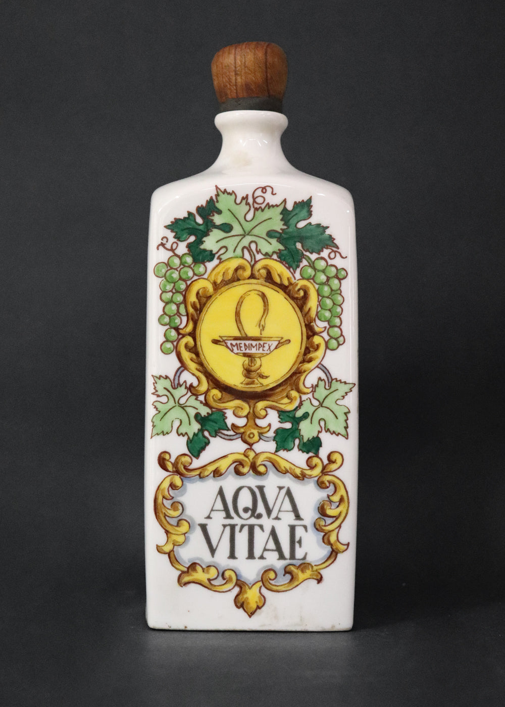 Vintage collectible Hollóházi porcelain bottle specially designed for "Aqua Vitae," which is an old Latin term for "water of life" typically referring to early distilled spirits. Medimpex, who’s name is on a medallion framed with scrolls was a well-known Hungarian/Eastern European pharmaceutical and medical distribution company. The bottle features a green and gold grapevine motifs framing a cartouche with the words "AQVA VITAE" boldly painted in the center. The wooden stopper is in perfect condition. Holló
