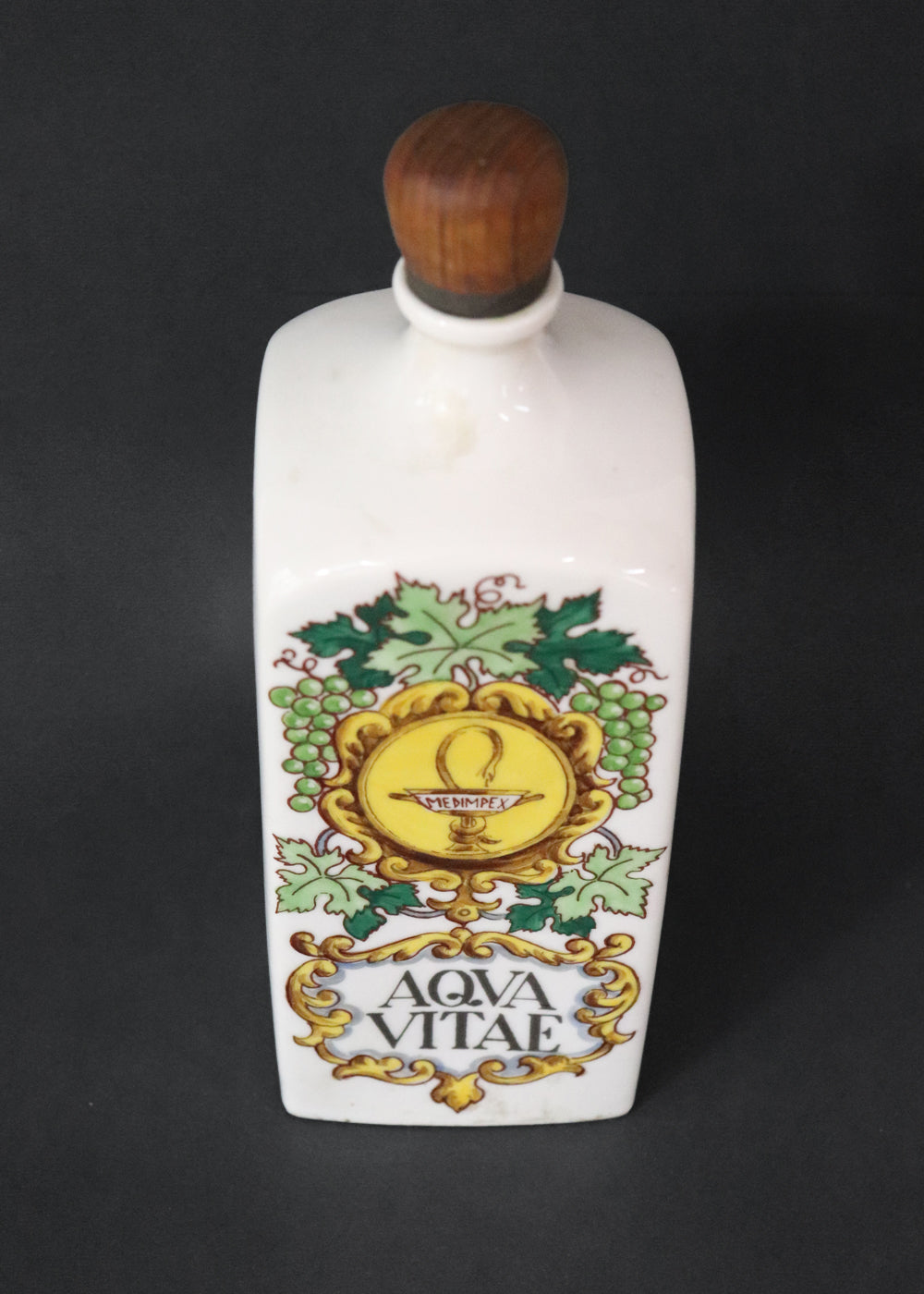 Vintage collectible Hollóházi porcelain bottle specially designed for "Aqua Vitae," which is an old Latin term for "water of life" typically referring to early distilled spirits. Medimpex, who’s name is on a medallion framed with scrolls was a well-known Hungarian/Eastern European pharmaceutical and medical distribution company. The bottle features a green and gold grapevine motifs framing a cartouche with the words "AQVA VITAE" boldly painted in the center. The wooden stopper is in perfect condition. Holló