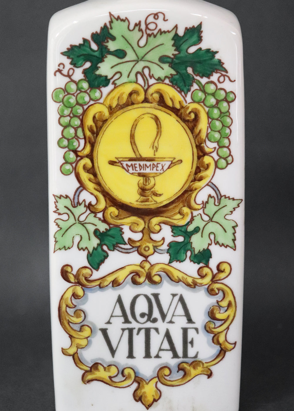Vintage collectible Hollóházi porcelain bottle specially designed for "Aqua Vitae," which is an old Latin term for "water of life" typically referring to early distilled spirits. Medimpex, who’s name is on a medallion framed with scrolls was a well-known Hungarian/Eastern European pharmaceutical and medical distribution company. The bottle features a green and gold grapevine motifs framing a cartouche with the words "AQVA VITAE" boldly painted in the center. The wooden stopper is in perfect condition. Holló
