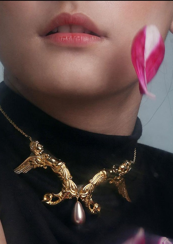 A gold-colored necklace with angel and mermaid design elements, featuring a shell pearl accent, displayed against a neutral background with a person wearing a black top and pink flower in the background.