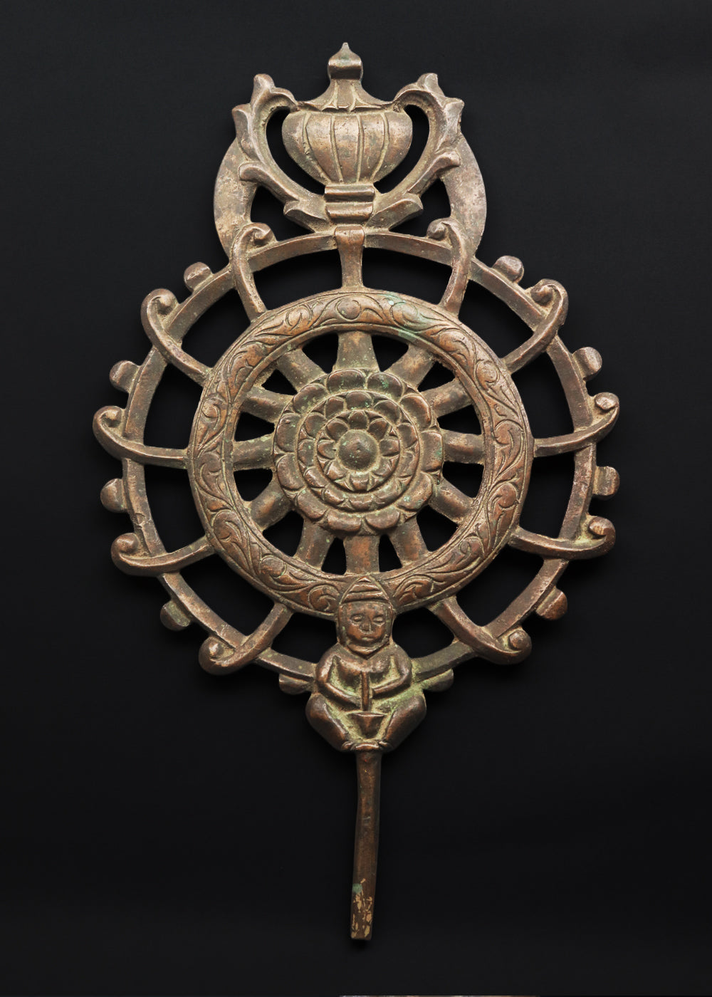 Antique cast bronze dharmachakra with a central lotus rosette within the spoked wheel. The top has an ornamental finial with a stupa like motif and the base features a devotional figure in a meditative pose holding an offering or bowl. The wavy, hook-like spokes projecting outward mimic flaming jewels or stylized rays common in Esoteric (Tantric) Buddhist art. A dharmachakra with 12 spokes symbolizes the Twelve Links of Dependent Origination (Pratītyasamutpāda), explaining the chain of cause and effect.