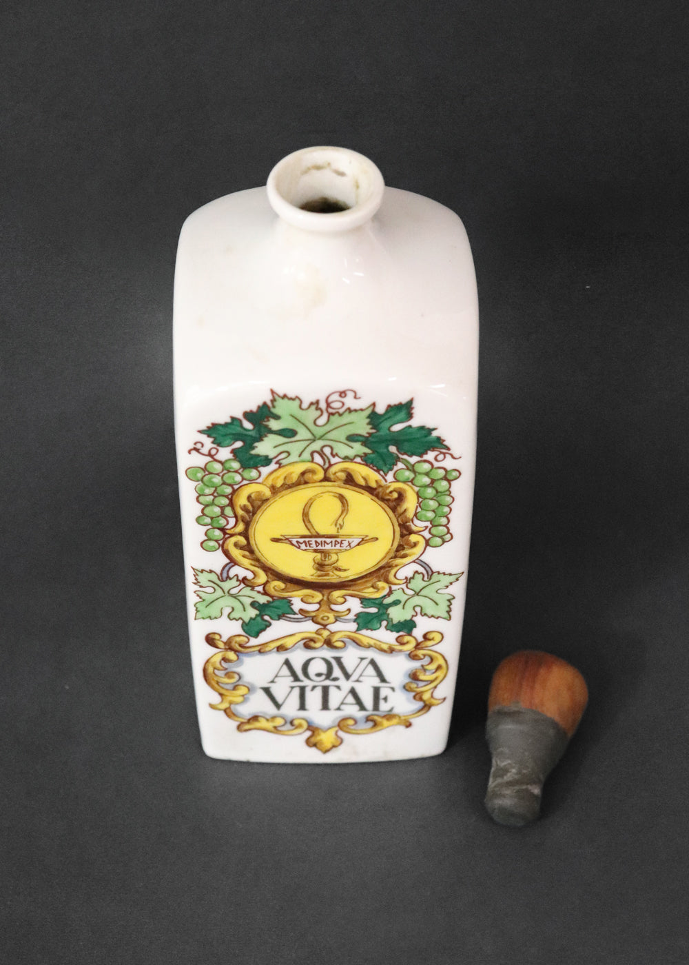 Vintage collectible Hollóházi porcelain bottle specially designed for "Aqua Vitae," which is an old Latin term for "water of life" typically referring to early distilled spirits. Medimpex, who’s name is on a medallion framed with scrolls was a well-known Hungarian/Eastern European pharmaceutical and medical distribution company. The bottle features a green and gold grapevine motifs framing a cartouche with the words "AQVA VITAE" boldly painted in the center. The wooden stopper is in perfect condition. Holló