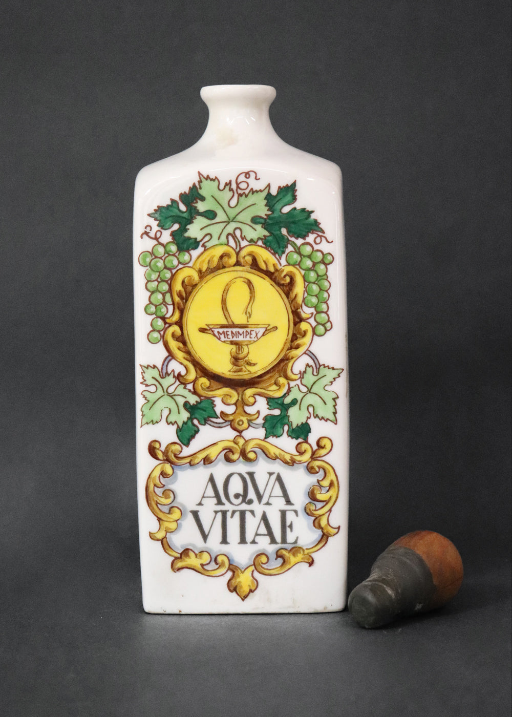 Vintage collectible Hollóházi porcelain bottle specially designed for "Aqua Vitae," which is an old Latin term for "water of life" typically referring to early distilled spirits. Medimpex, who’s name is on a medallion framed with scrolls was a well-known Hungarian/Eastern European pharmaceutical and medical distribution company. The bottle features a green and gold grapevine motifs framing a cartouche with the words "AQVA VITAE" boldly painted in the center. The wooden stopper is in perfect condition. Holló
