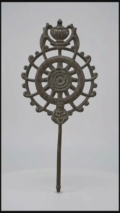Antique cast bronze dharmachakra with a central lotus rosette within the spoked wheel. The top has an ornamental finial with a stupa like motif and the base features a devotional figure in a meditative pose holding an offering or bowl. The wavy, hook-like spokes projecting outward mimic flaming jewels or stylized rays common in Esoteric (Tantric) Buddhist art. A dharmachakra with 12 spokes symbolizes the Twelve Links of Dependent Origination (Pratītyasamutpāda), explaining the chain of cause and effect.