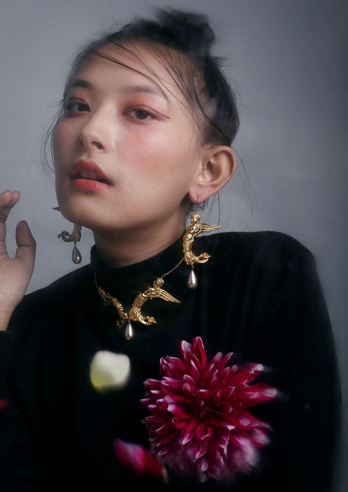 A person wearing half mermaid, half angel earrings with gold plating and champagne colored shell pearl accents, with a floral pattern in the background.